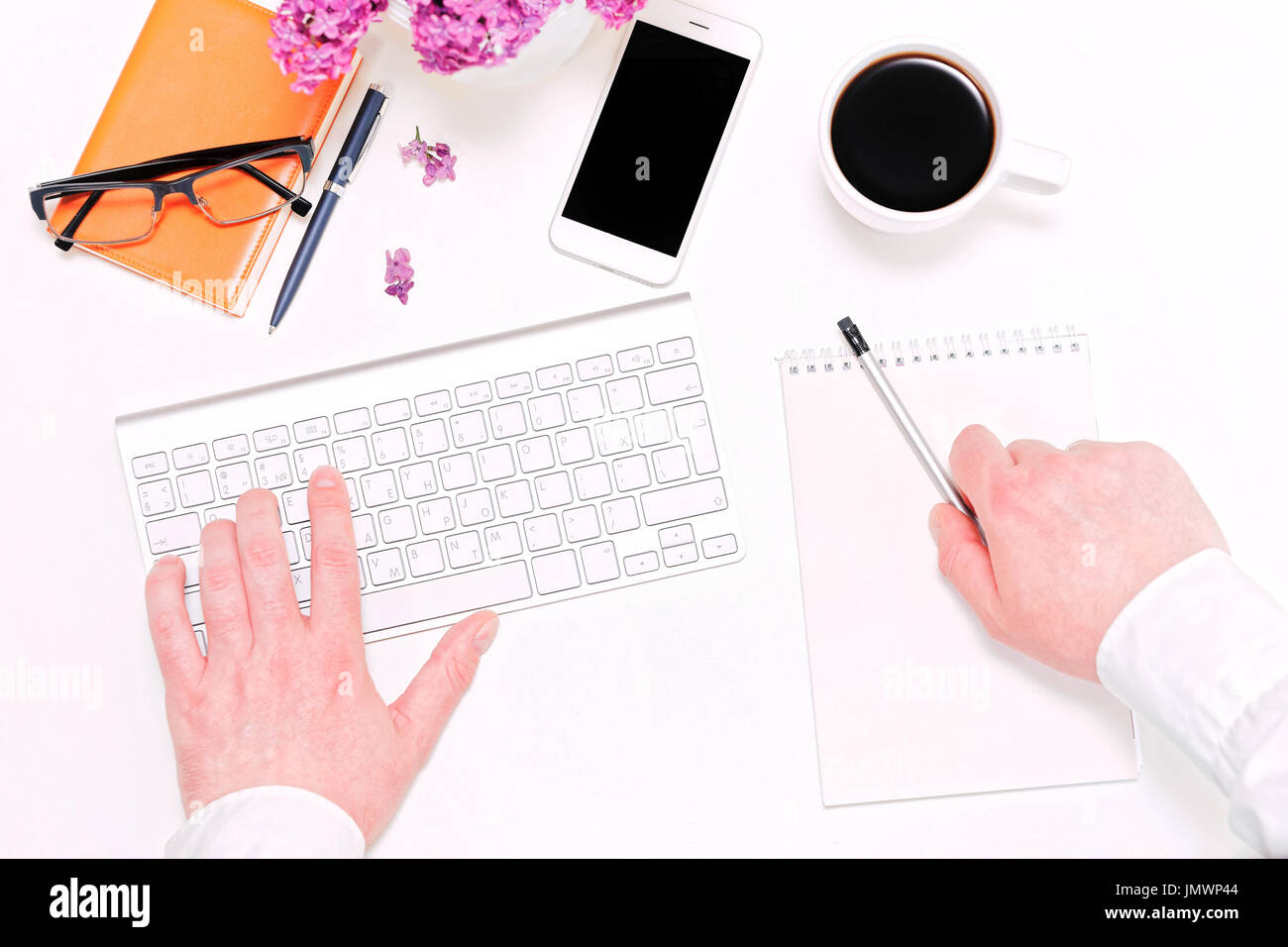 Mann schreiben Notizen im Tagebuch. Arbeitsplatz mit Computer-Tastatur, Handy, Tasse Kaffee, Notizbuch, Kugelschreiber, Bleistift, Gläser und lila Blumen auf der whi Stockfoto