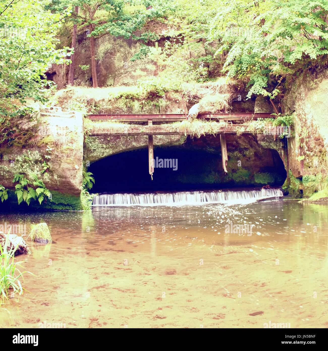 Kleinen Wehr am Fluss fließt aus der Höhle. Kaltes Wasser des kleinen Fluss über kleine steinerne Wehr. Steinige und rostigen Konstruktion. Stockfoto