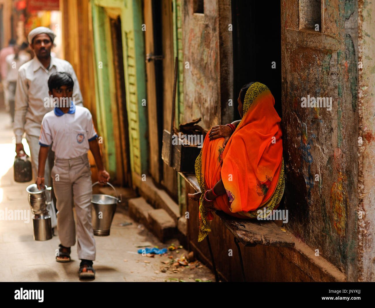 Eine Frau in einem hellen orange Sari sitzt außerhalb ihrer Heimat als junge und Mann vorbei, Edelstahl Behälter Stockfoto