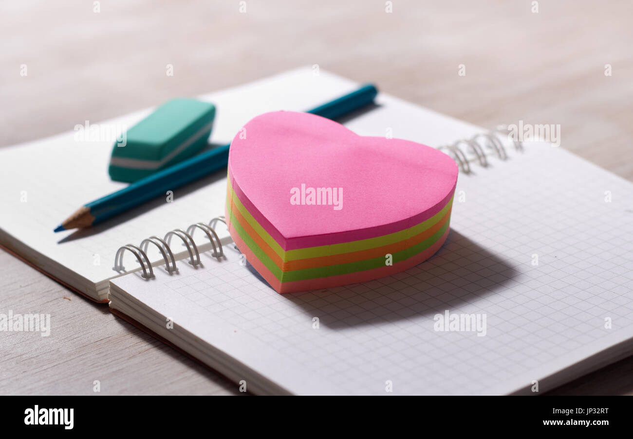 Herzförmige Aufkleber auf leeren Notizbuch mit Bleistift und Radiergummi im Hintergrund, am Schreibtisch aus Holz Stockfoto