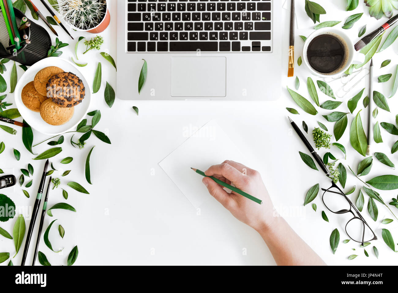 Teilweisen Draufsicht der Person halten Sie Stift und schreiben am Arbeitsplatz mit Laptop, Tasse Kaffee, grünen Blättern und Bürobedarf, drahtlose communicatio Stockfoto