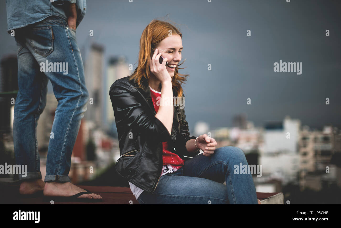 Eine Frau sitzen und reden über ein Mobiltelefon auf eine Stadt auf dem Dach, mit einem Mann hinter ihr stehen. Stockfoto