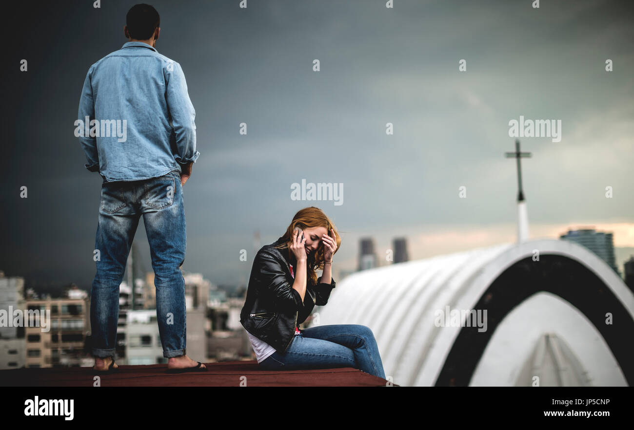 Eine Frau sitzen und reden über ein Mobiltelefon auf eine Stadt auf dem Dach, mit einem Mann hinter ihr stehen. Stockfoto