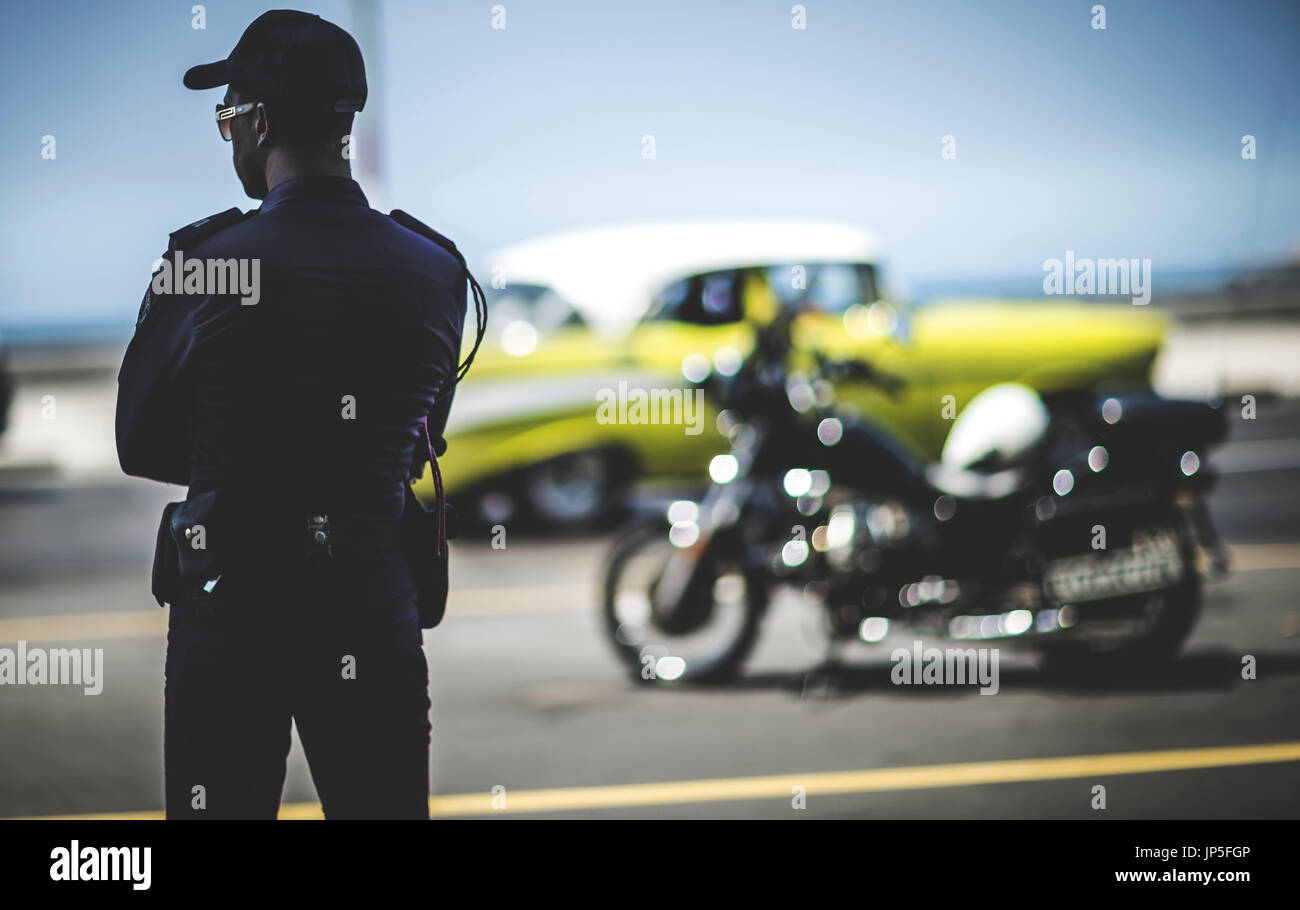 Rückansicht des Mannes in Uniform mit Radio und Waffe Gürtel stehen mit Blick auf einen Parkplatz. Motorrad und Oldtimer im Hintergrund. Stockfoto