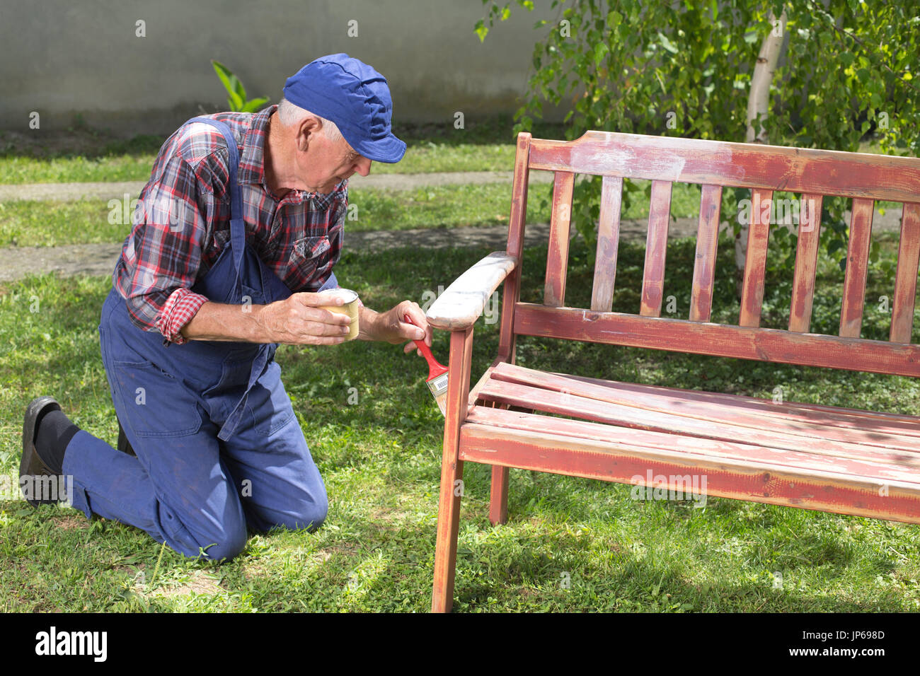 Senior Mann im Overall Malerei alte Bank im Garten nach dem Sandstrahlen. Reparatur von alten Möbeln Stockfoto