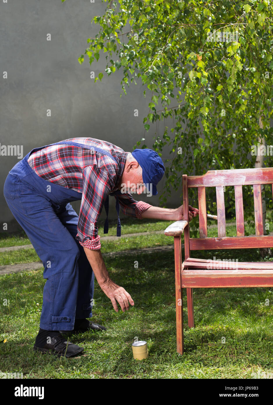 Senior Mann im Overall Malerei alte Bank im Garten nach dem Sandstrahlen. Reparatur von alten Möbeln Stockfoto