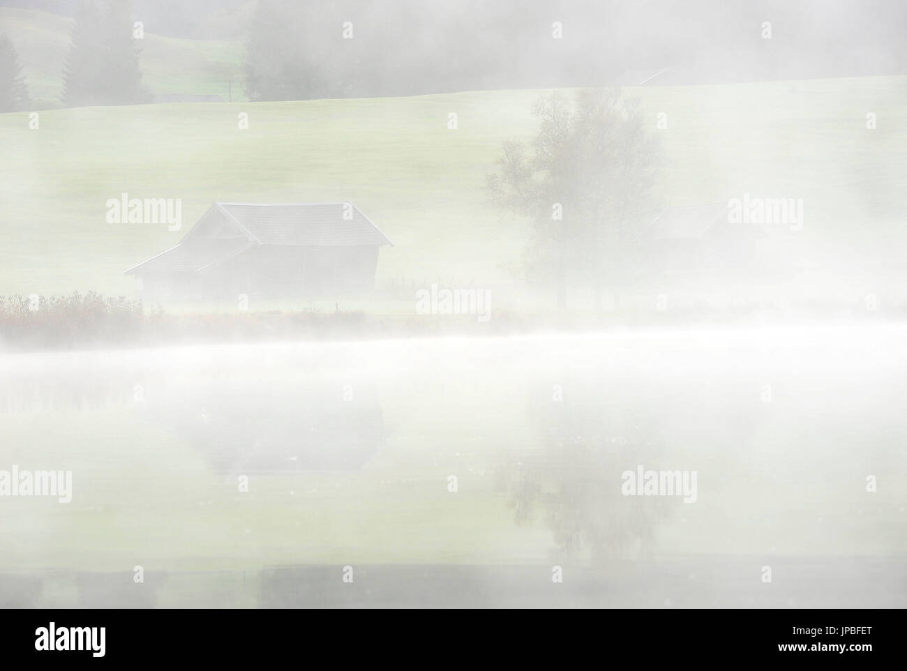 Spiegelung von einem Heuhaufen und Baum in das Wasser des kleinen Alpensee mit Nebel. Stockfoto