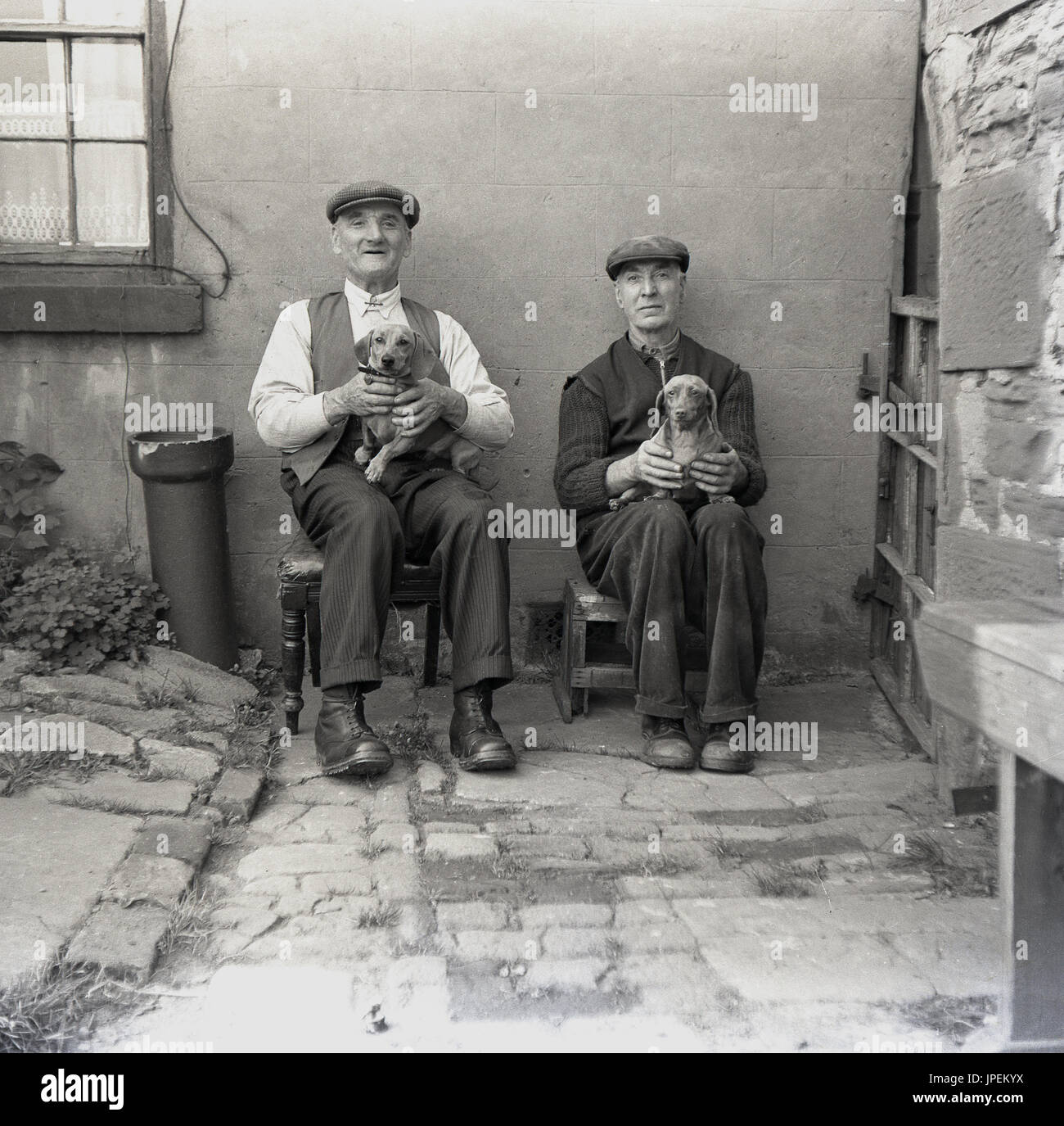 draußen Sie 1940er Jahre, England, Lancaster, zwei ältere nördliche Männer tragen Schiebermütze und Stiefel, in einem Hof auf kleine Stände mit Stolz ihre kleinen Hunde halten. Stockfoto