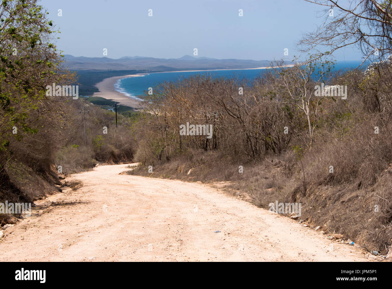Eine unbefestigte Landstraße führt bergab durch trockenen Busch mit einem langen Strand und das Meer im Hintergrund. Stockfoto