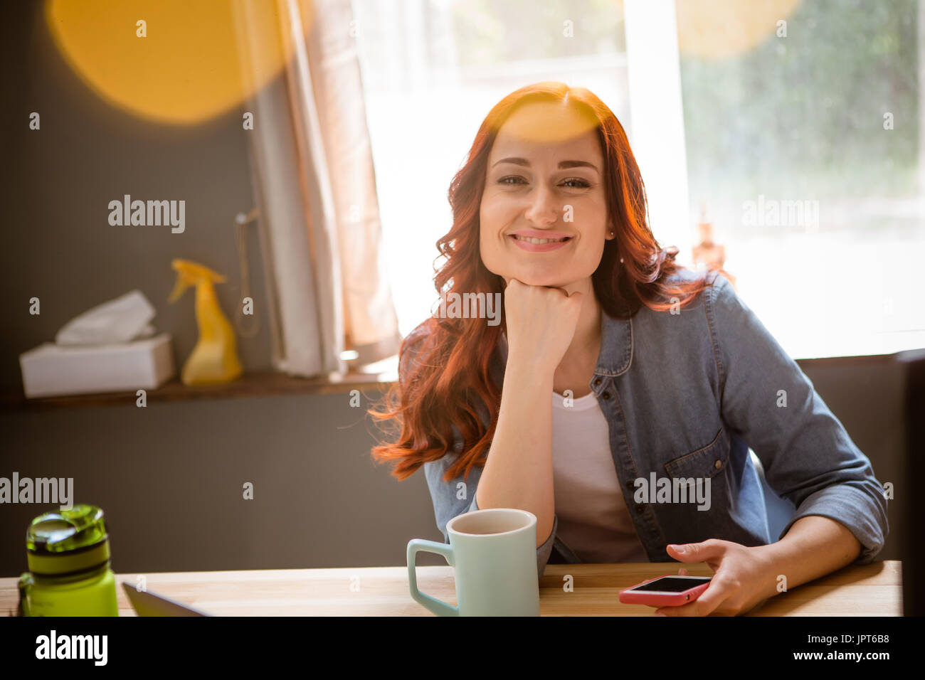 Porträt von foxy Frau arbeitet auf Frelance aus der Ferne. Frau zu Hause im Büro sitzen am Tisch mit Hand schließen um Kinn, lächelnd. Getönten Bild mit Sonne Stockfoto