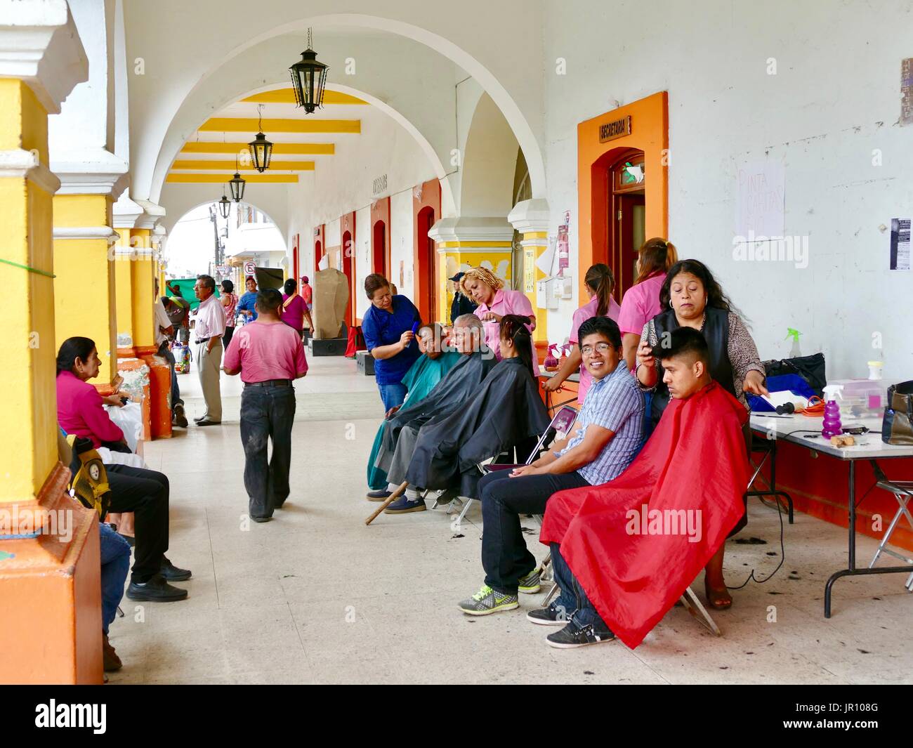Männer aller Altersgruppen, eine Frau, die Bewertungsabschläge, die im freien Platz mit Arkaden. Junge Mann in die Kamera lächelt. Friseure arbeiten. Zaachila, Oaxaca, Mexiko. Stockfoto