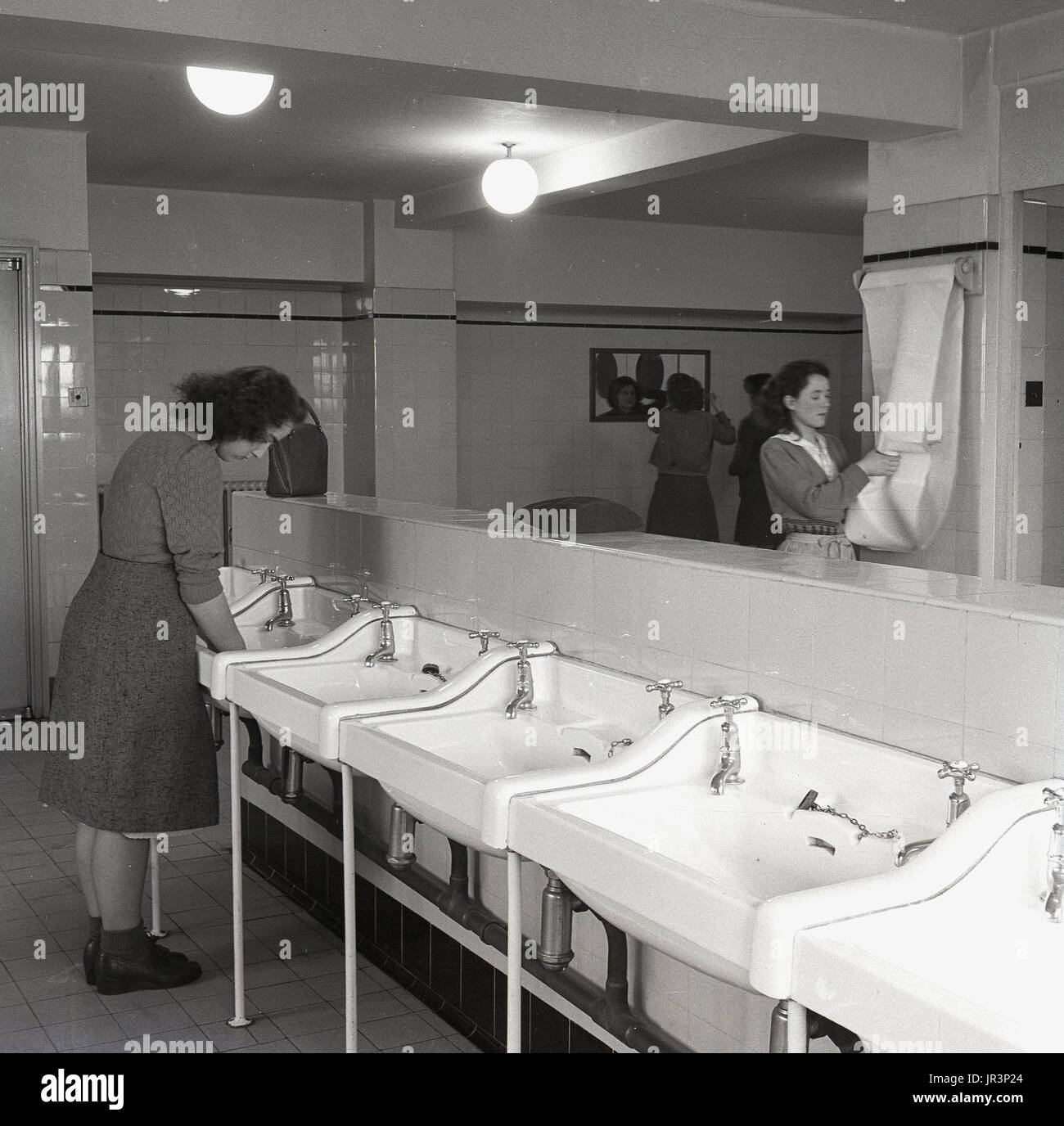 1950, England, junge erwachsene Weibchen Reinigung ihre Hände in großen Waschbecken und ihr Aussehen in einem Spiegel Kontrolle in einem gefliesten Badezimmer an einem Teachers Training College. Stockfoto