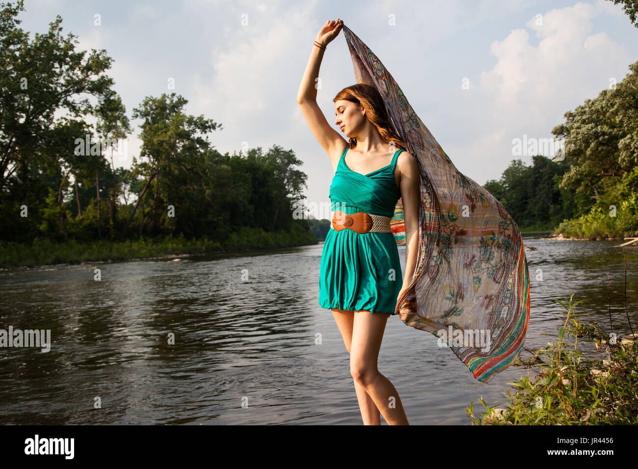 Junge Frau mit einem Kleid Petrol Grün steht in der Nähe von einem Fluss und See hält Teal Schal im Wind.   Sie ist entspannt und gelassen, während das Gefühl der Sommer Stockfoto