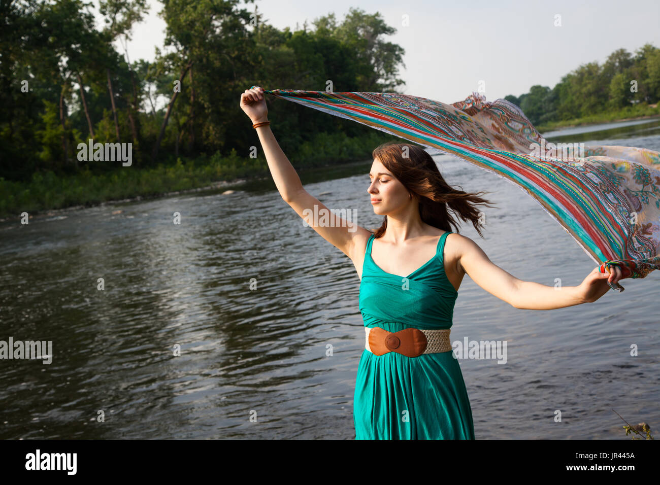 Junge Frau mit einem Kleid Petrol Grün steht in der Nähe von einem Fluss und See/Petrol bunten Schal in den Wind hält.   Sie ist entspannt und heiter, während das Gefühl th Stockfoto