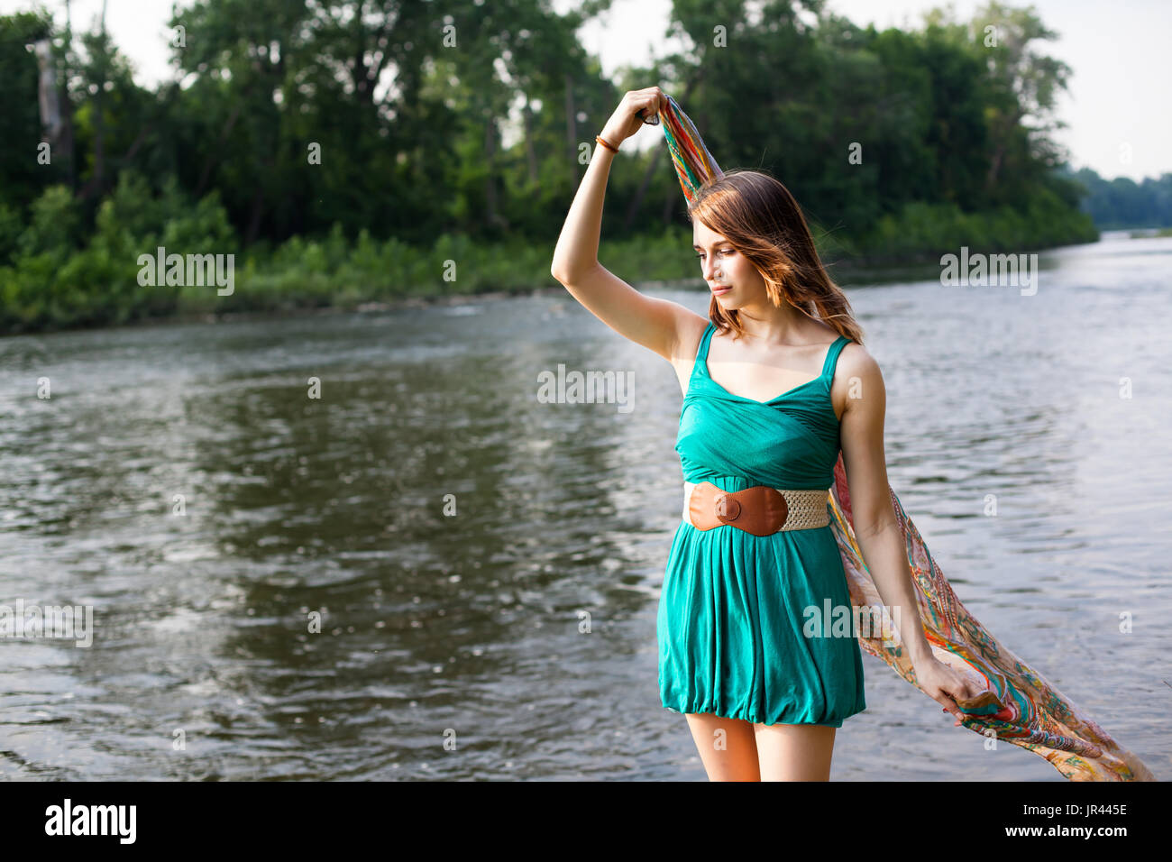 Junge Frau mit einem blaugrün Kleid steht in der Nähe von einem Fluss und See hält Teal Schal im Wind.   Sie ist entspannt und ruhig während der Sommer Brise Stockfoto