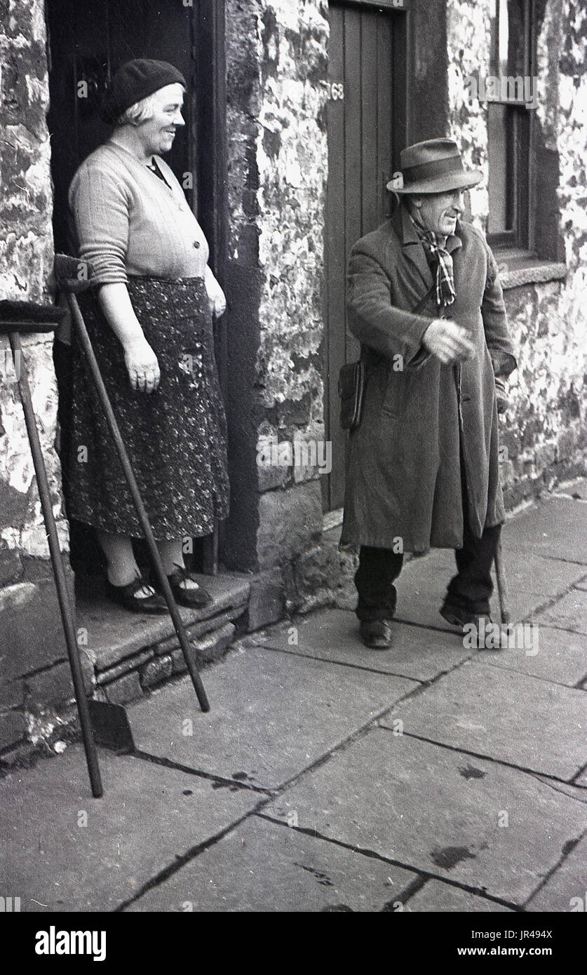 1940 s, historischen, älteren Herrn trug einen Mantel und Hut und Dame in Barett am Eingang zu Ihrem Ferienhaus aus Stein, Merthyr Tydfil, South Wales, UK. Der Mann versucht, Herzmuscheln er am Strand von Tür zu Tür bezogen hat, verkaufen. Stockfoto