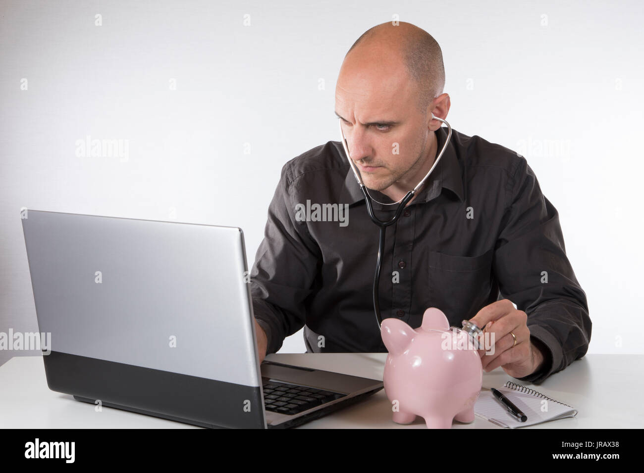 Geschäftsmann in der finanziellen Mühe Fehlerbehebung seine kleine rosa Sparschwein mit einem Stethoskop, während er an seinem Computer in einem konzeptionellen Image Works Stockfoto