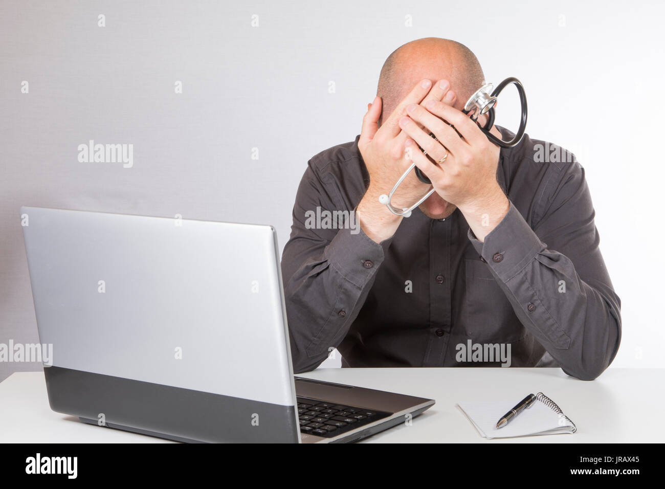 Verzweifelt oder der Arzt seinen Kopf in seine Hände erschöpft, wie er sitzt an seinem Schreibtisch arbeiten auf einem Laptop. Stockfoto