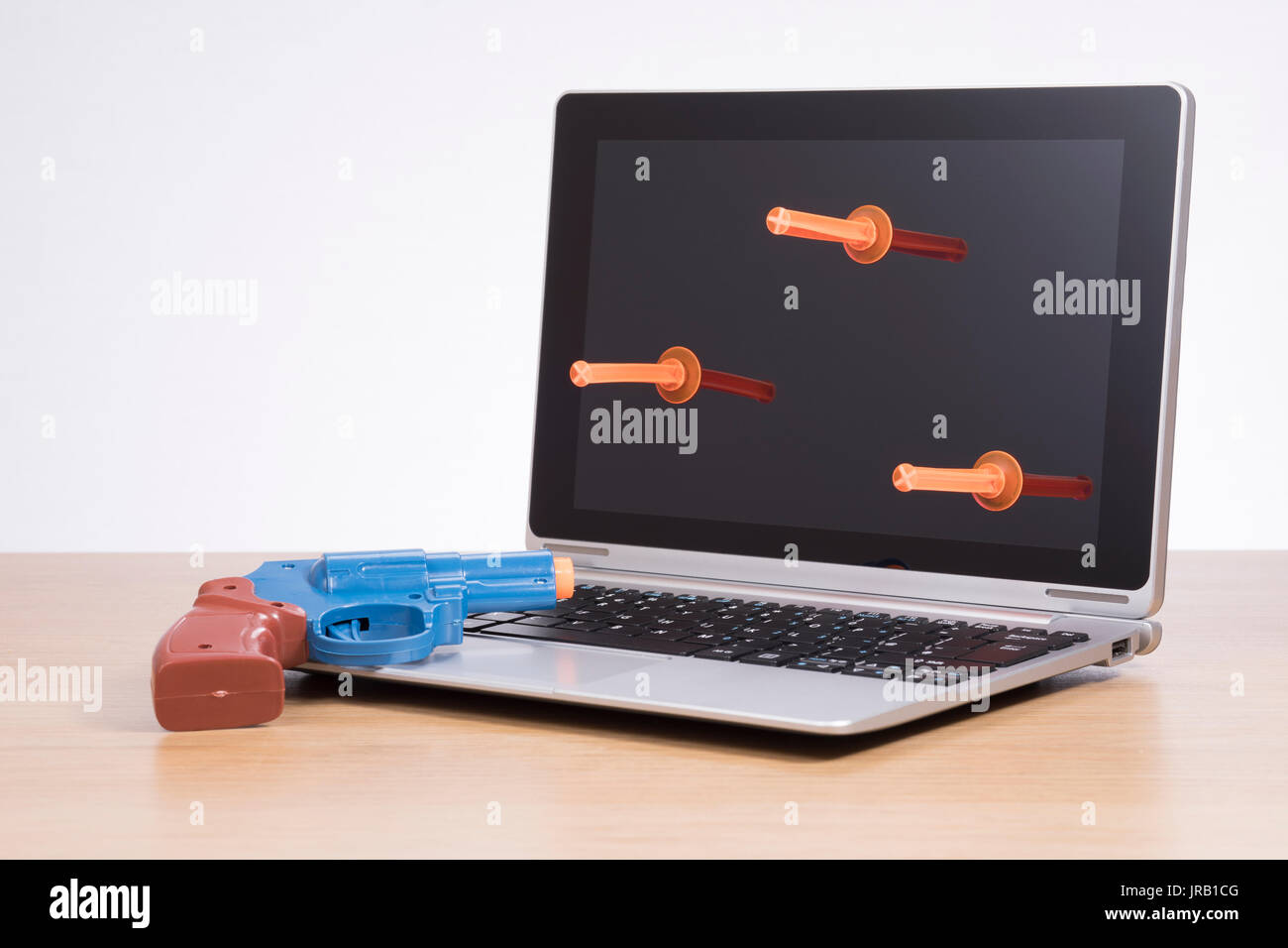 Ein Spielzeug, Kunststoff Pistole ruht auf der Tastatur eines Laptops auf einem Holz desk top Wie drei Sog Darts aus der leeren, schwarzen Bildschirm hängen. Stockfoto