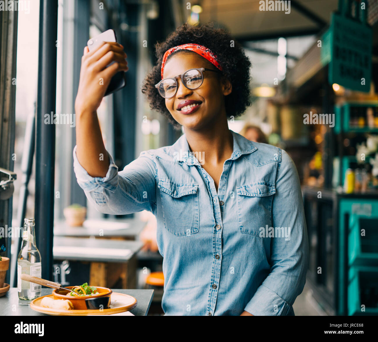 Attraktive junge afrikanische Frau lächelnd und selfie mit Ihrem Smartphone, während allein in einem Café genießen Sie eine Mahlzeit sitzen Stockfoto