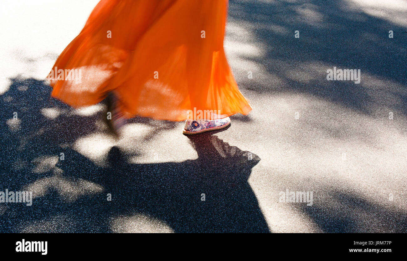 Beine einer jungen Frau zu Fuß in langen orangefarbenen Kleid in summer breeze Flattern mit Bürgersteig Baum Schatten Stockfoto