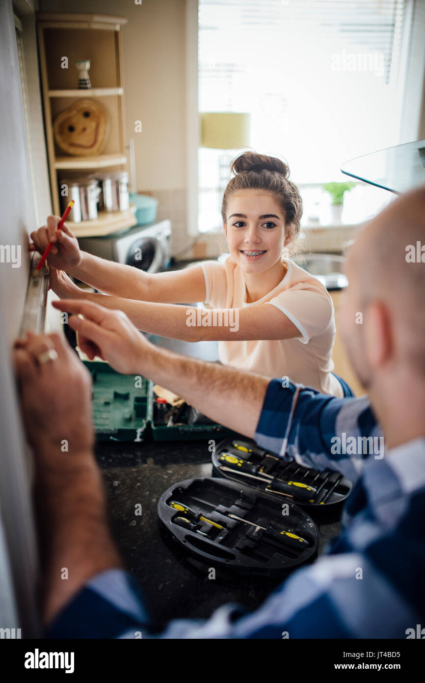 Teenage Mädchen ihren Vater mit DIy zu helfen in Ihrer Küche. Das Mädchen ist die Kennzeichnung der Wand mit einem Bleistift, während ihr Vater lächelnd. Stockfoto