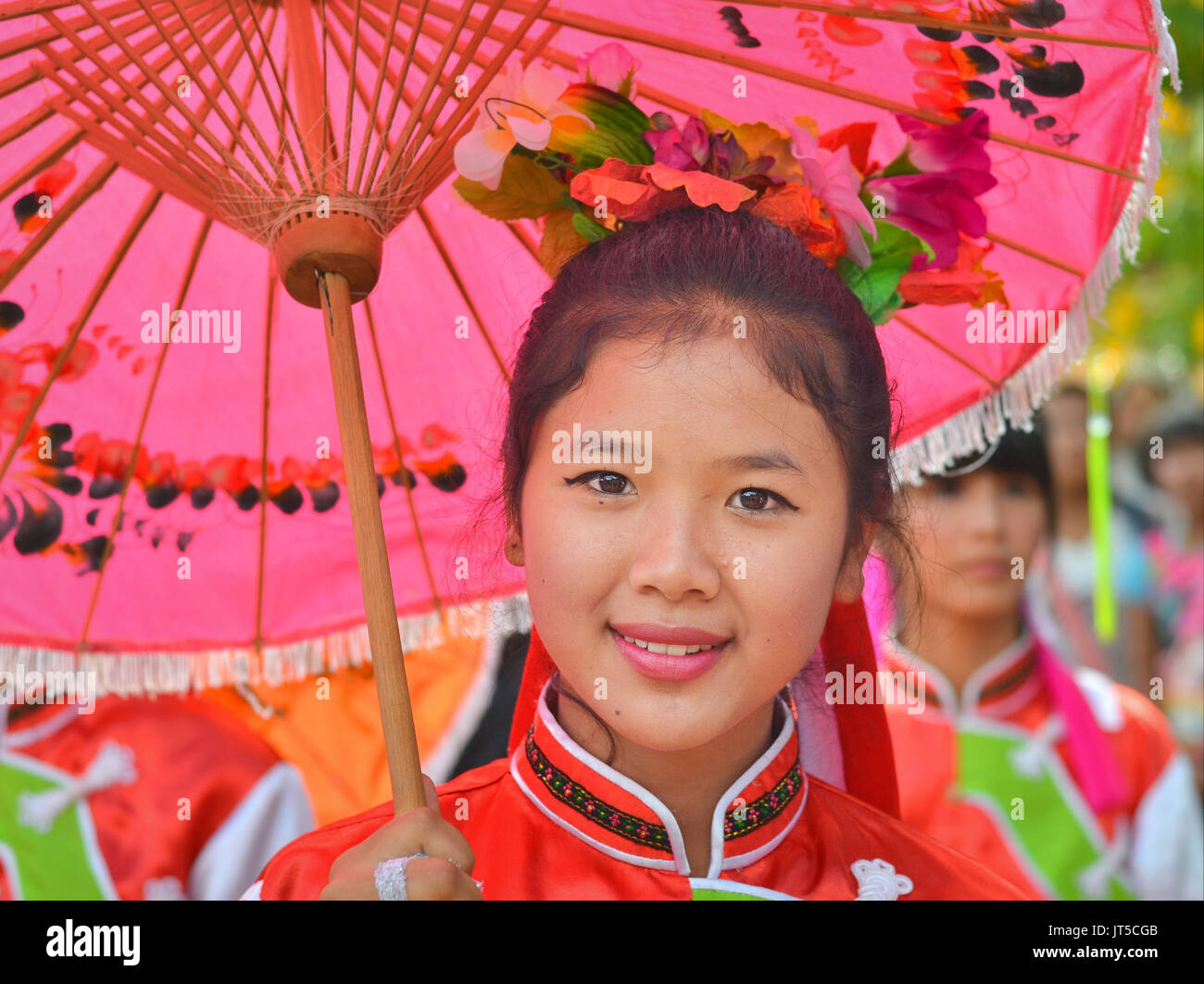 Hübsches thailändisches chinesisches Mädchen im traditionellen Kleid mit rotem Sonnenschirm lächelt für die Kamera. Stockfoto
