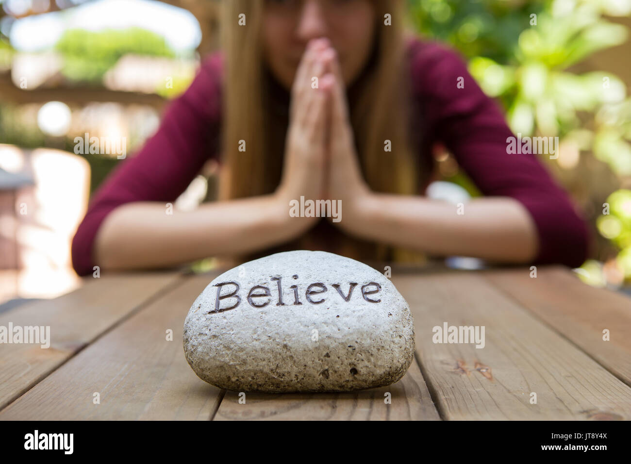 Junge Frau sitzen und Beten an einem Holztisch im Hintergrund mit einem Rock, der vor ihr mit dem Wort bezeichnet Glauben. Model Release. Stockfoto