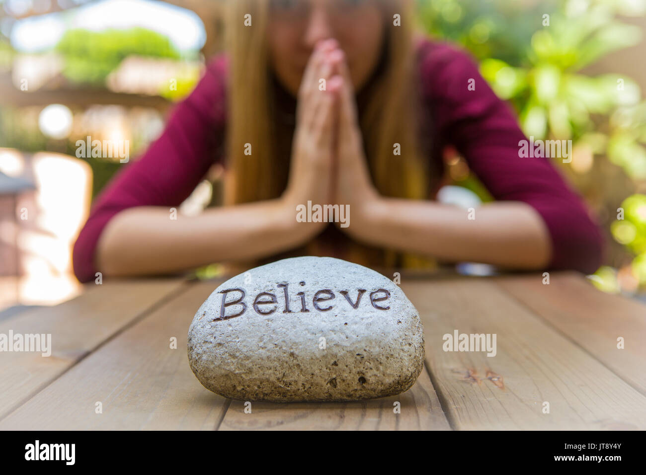 Junge Frau sitzen und Beten an einem Holztisch im Hintergrund mit einem Rock, der vor ihr mit dem Wort bezeichnet Glauben. Model Release. Stockfoto