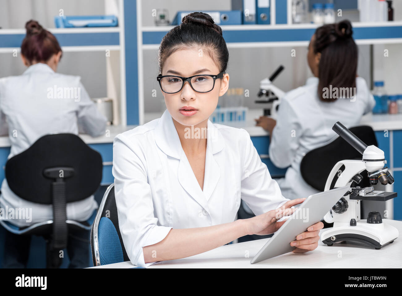 Asiatische Wissenschaftler in Laborkittel mit Mikroskop und digitale Tablette im chemischen Labor Wissenschaftler hinter Gruppe Stockfoto