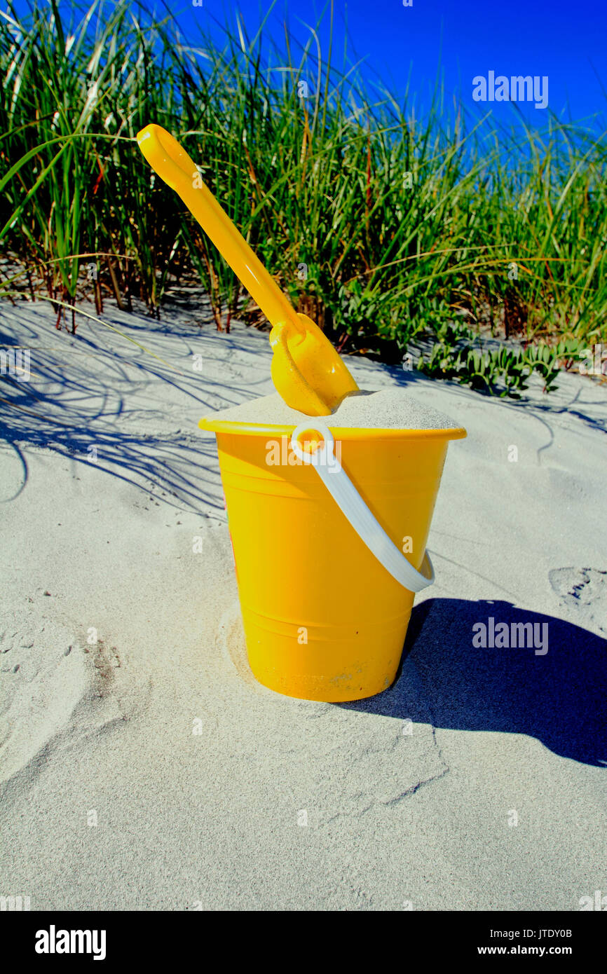 Ein Kind im gelben Eimer mit Sand und mit einer Schaufel an einem Strand gefüllt Stockfoto