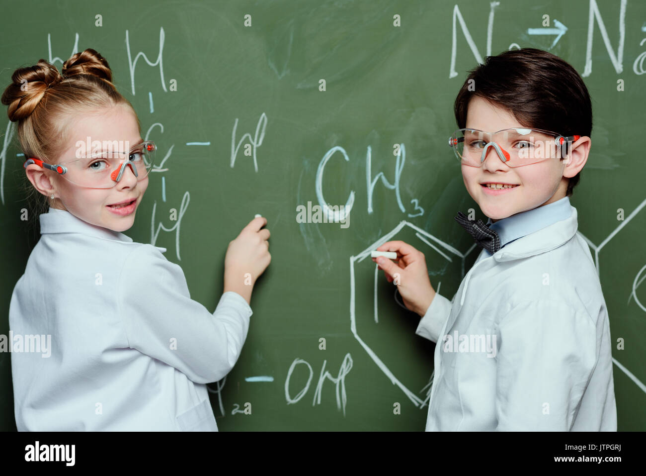 Schüler im weißen Kittel und Schutzbrille Zeichnen chemischer Formeln auf der Schiefertafel und Kamera, Wissenschaft Schule Konzept Stockfoto