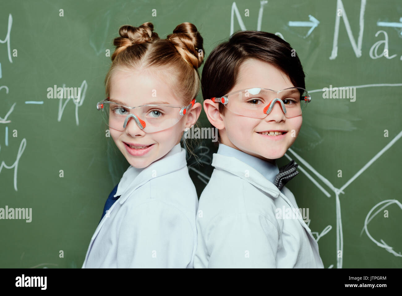 Jungen und Mädchen im weißen Kittel und Schutzbrille zusammen in der Nähe der Tafel stand und lächelte Kamera, Wissenschaft Schule Konzept Stockfoto