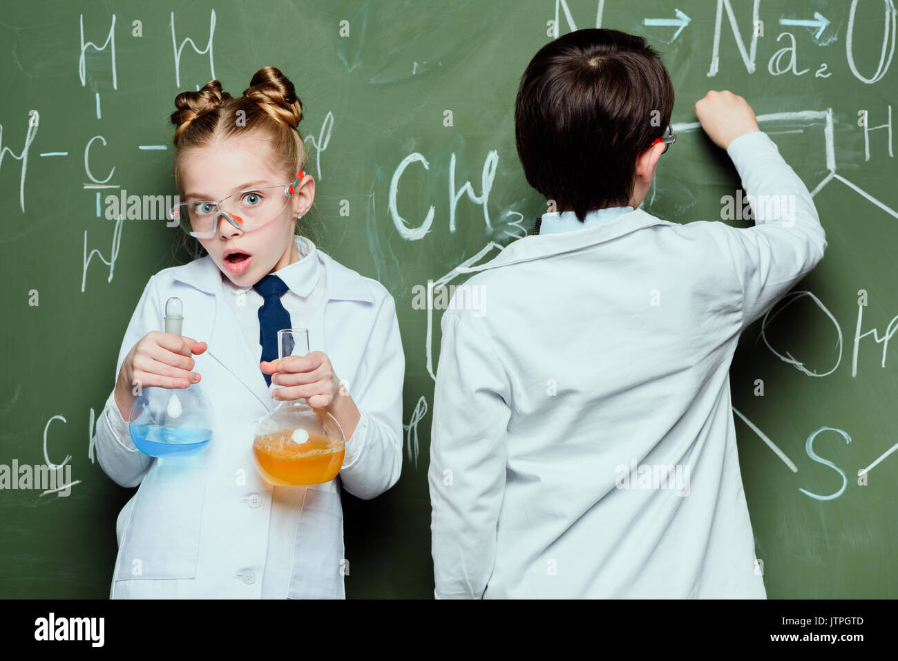 Junge und Mädchen in weißen Kitteln mit Reagenzien in Flaschen und Zeichnen chemischer Formeln in der Wissenschaft Labor Stockfoto