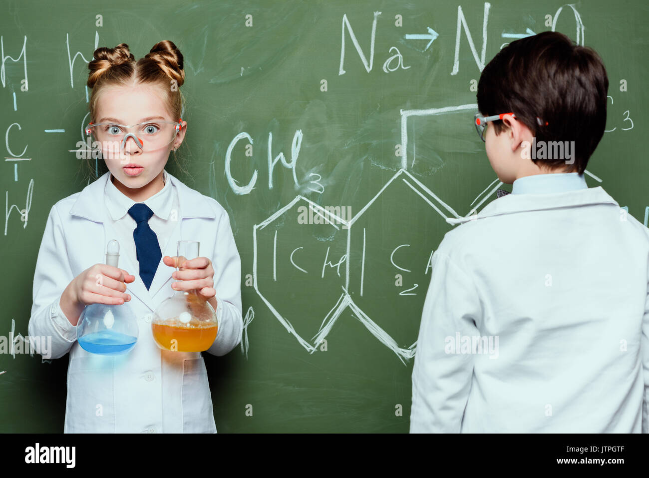 Junge und Mädchen in weißen Kitteln mit Reagenzien in Flaschen und Zeichnen chemischer Formeln in der Wissenschaft Labor Stockfoto