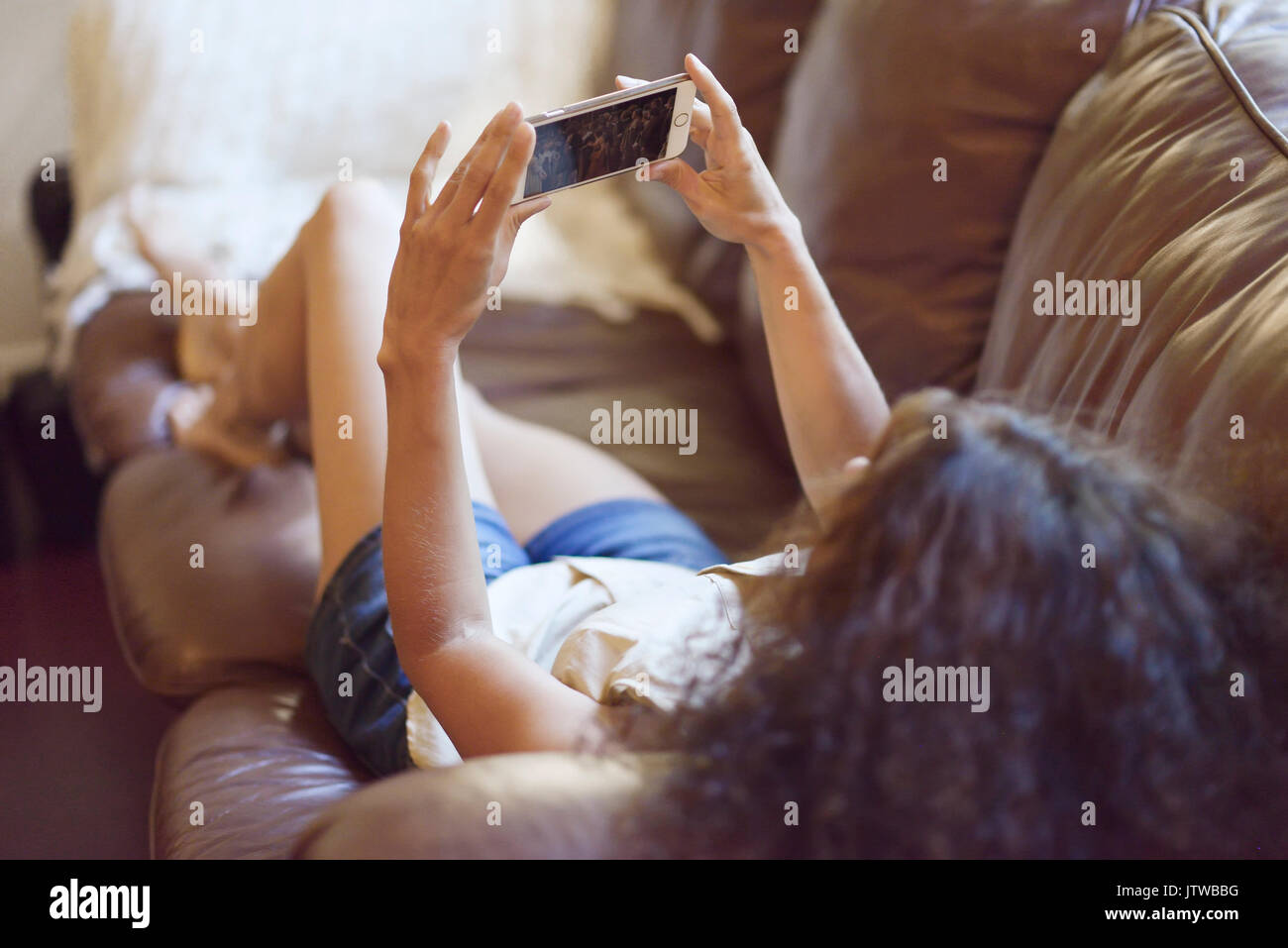 Junge Frau auf einem Sofa zu Hause liegen mit einem iPhone in den Händen, einen Film auf dem Bildschirm "Telefon" Stockfoto