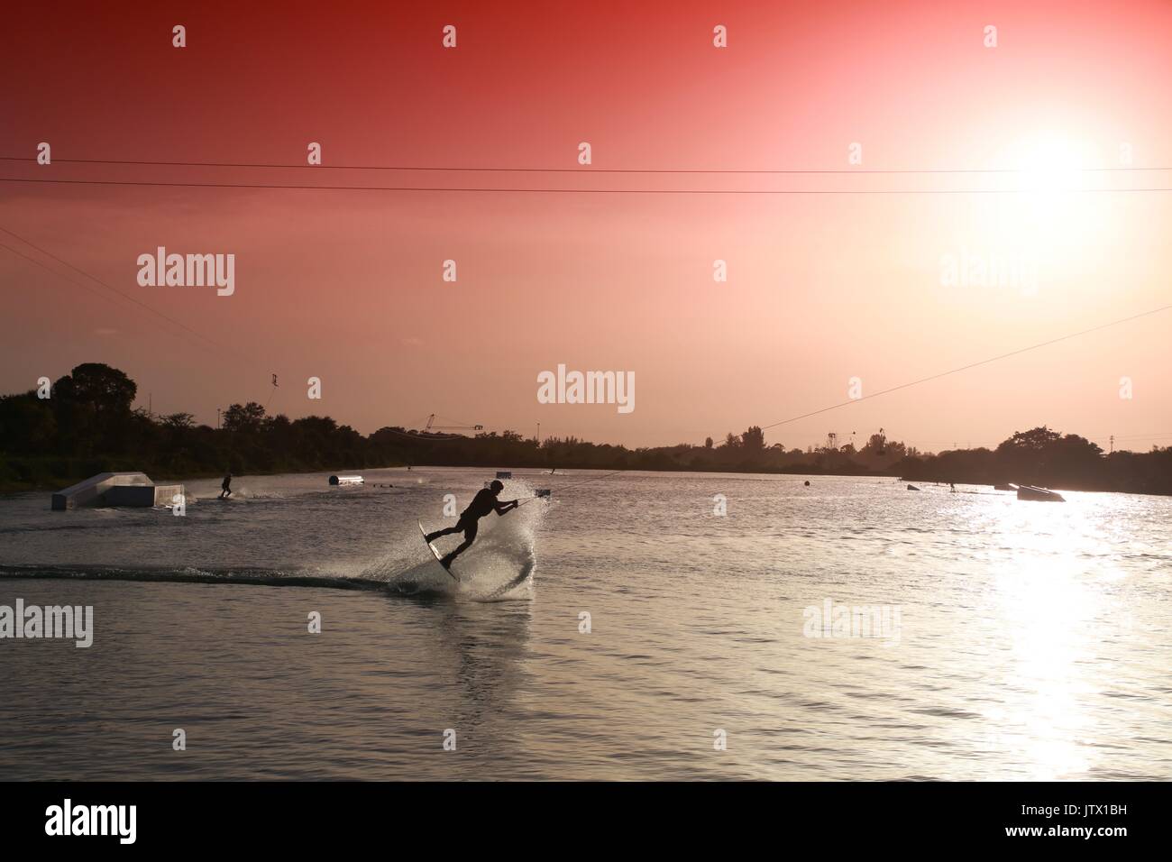 Wake Boarder in Mid-Air tun Half-Somersault auf dem See bei Sonnenuntergang hinter Plume von Spritzwasser durch Rot abgestufte Filter Sky akzentuieren Stockfoto