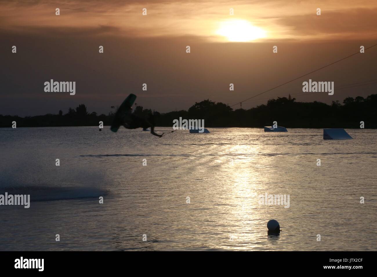 Wake Boarder in Mid-Air tun Half-Somersault auf dem See bei Sonnenuntergang hinter Plume von Spritzwasser durch Braun graduiert Filter Sky akzentuieren Stockfoto