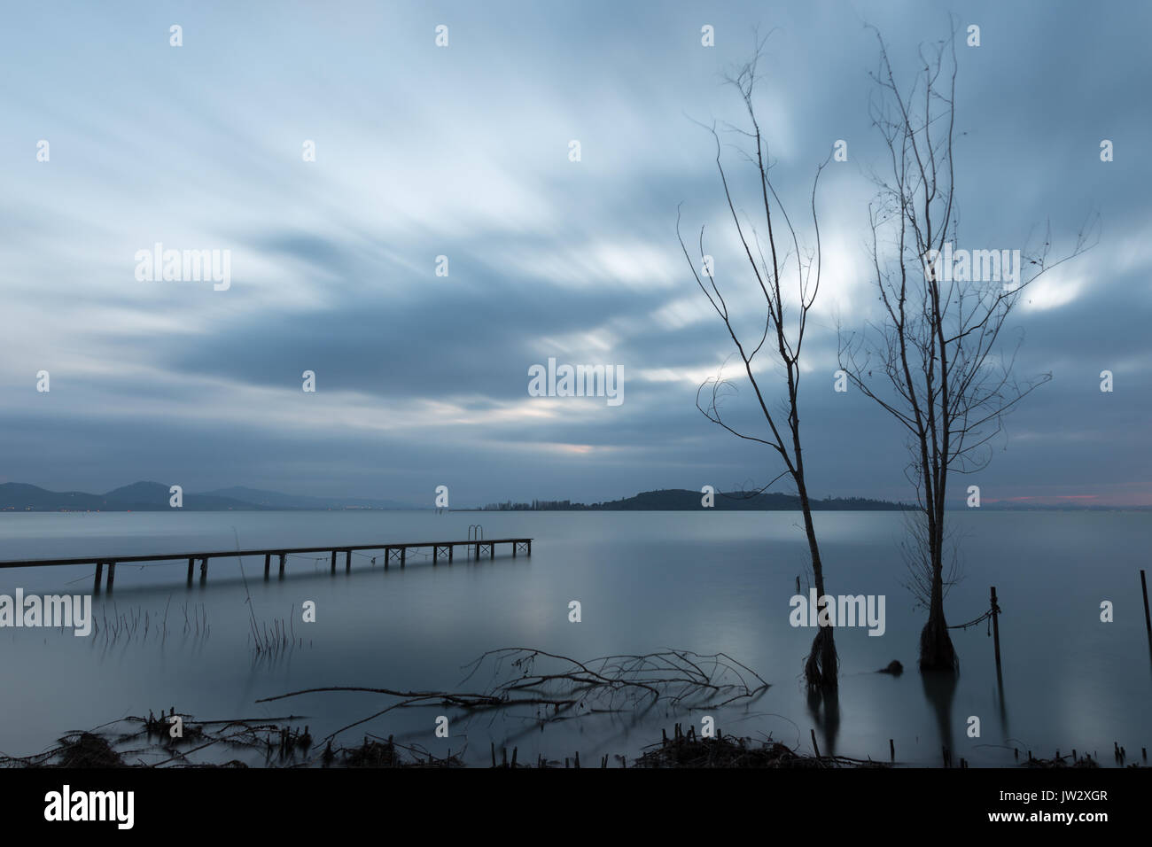 Einige der Skelettmuskulatur Bäume und ein Pier an einem See in der Abenddämmerung, mit perfekt noch Wasser und ziehenden Wolken Stockfoto