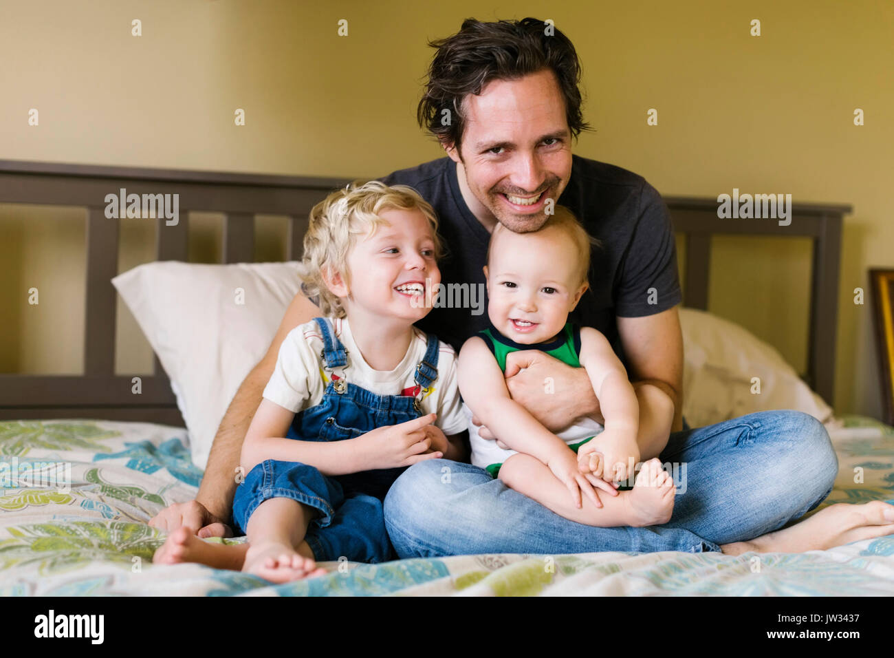 Vater mit Söhnen (12-17 Monate, 4-5) sitzen auf dem Bett Stockfoto