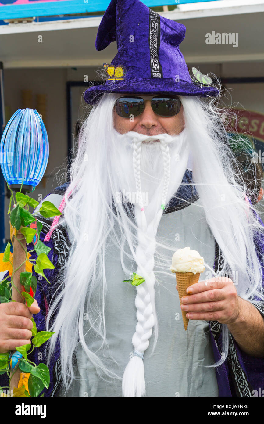 Burley, Hampshire, UK. 12 Aug, 2017. New Forest Fairy Festival. Massen von Feen und Elfen, mit Feenstaub besprüht, auf Burley für das Wochenende steigen für eine magische zauberhafte Festival im New Forest. Mann verkleidet als Zauberer in Mantel und Hut mit langen weißen Haar und Bart, in der einen Hand und Eis in der anderen abzuwehren. Credit: Carolyn Jenkins/Alamy leben Nachrichten Stockfoto