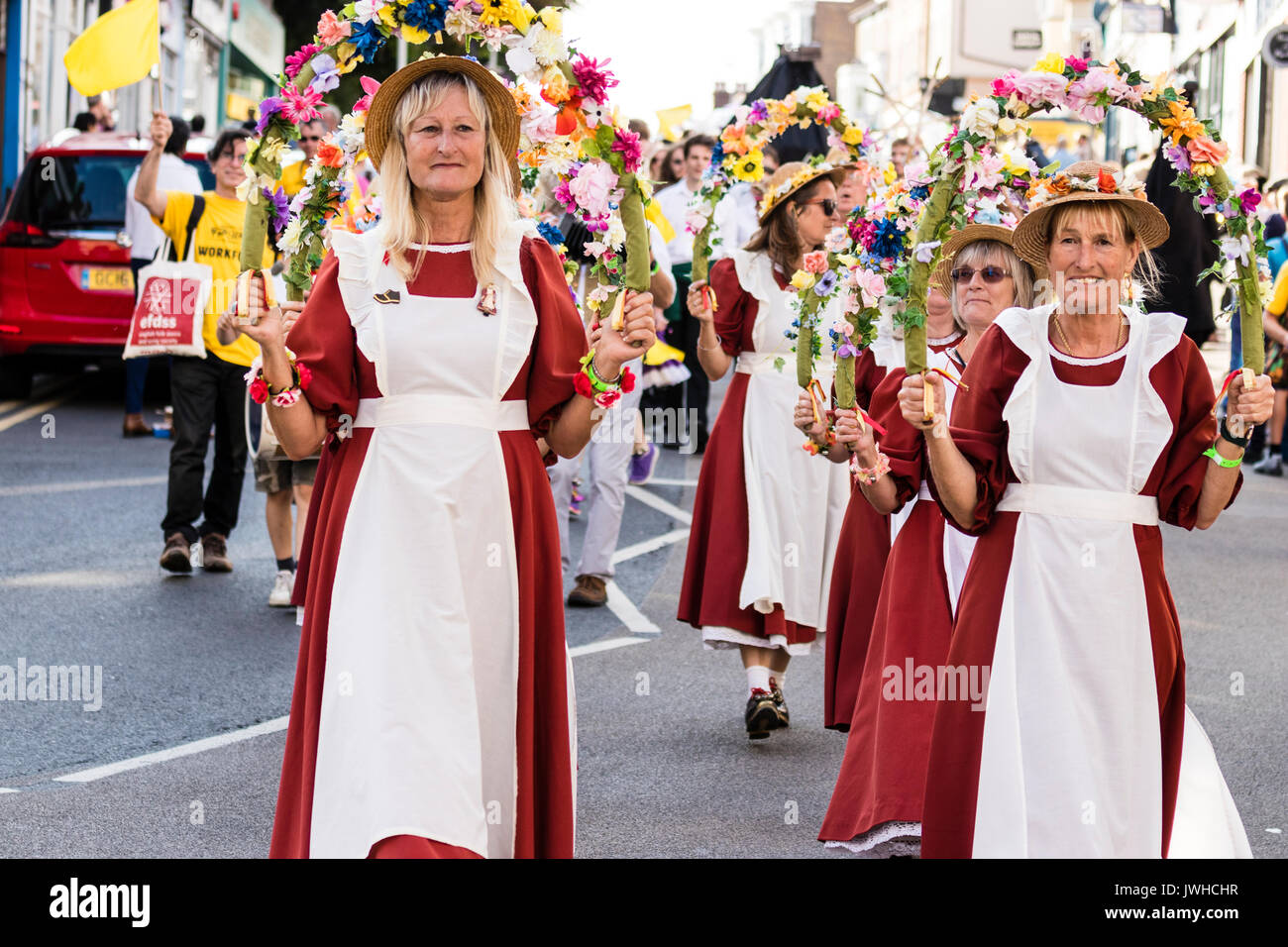 Broadstairs Folk Woche Festival Parade. Volkstänzer, der steigende Lerchen Frauen Morris Team, marschieren entlang der High Street, halten die Hälfte Girlanden aus Blumen über die Köpfe. Tragen orange Kleidern mit weißen Schürzen und Strohhüte. Stockfoto