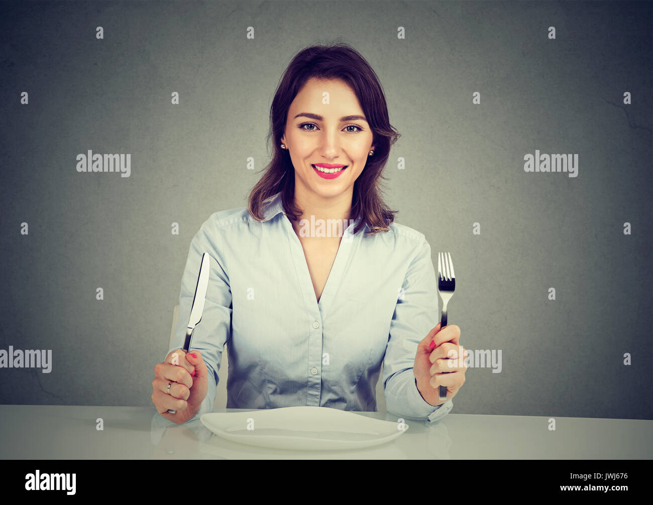 Glückliche Frau mit Gabel und Messer mit leeren Teller auf graue Wand Hintergrund isoliert am Tisch sitzen Stockfoto