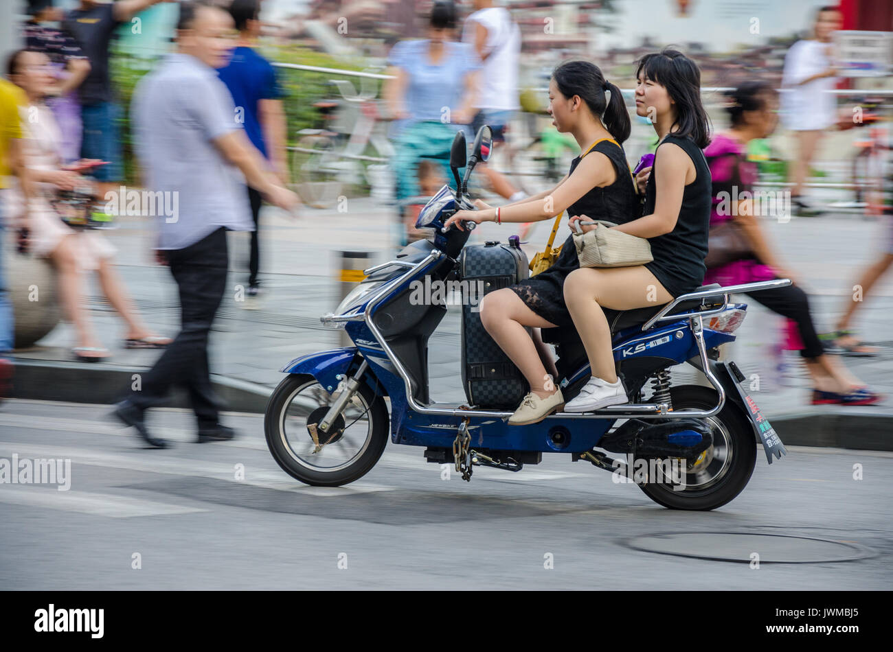 Zwei chinesische Damen auf einen elektrischen Moped style Roller Ansatz einen Fußgängerüberweg. Der Treiber hat einen Koffer zwischen die Beine. Stockfoto