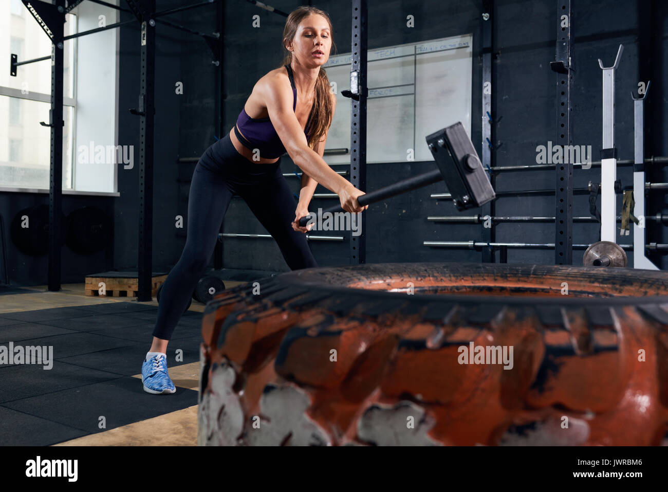 Portrait von starken jungen Frau smashing große Reifen mit vorschlaghammer während crossfit Workout im modernen Fitnessraum Stockfoto