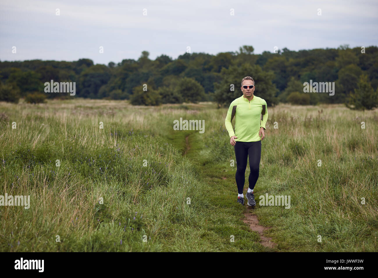Passen muskulösen Mann joggen auf einem ländlichen Trail durch Grünland trägt Sportkleidung und Sonnenbrille in einem aktiven Lifestyle Konzept Stockfoto