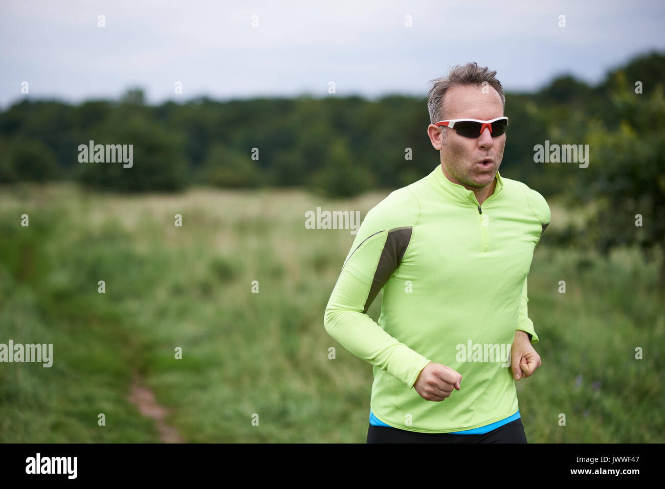 Gesund fit Mann mit Sonnenbrille Joggen durch ländliche Grünland auf einer Country Track mit Kopie Raum hinter ihm auf einem unscharfen Hintergrund in Nahaufnahme Stockfoto