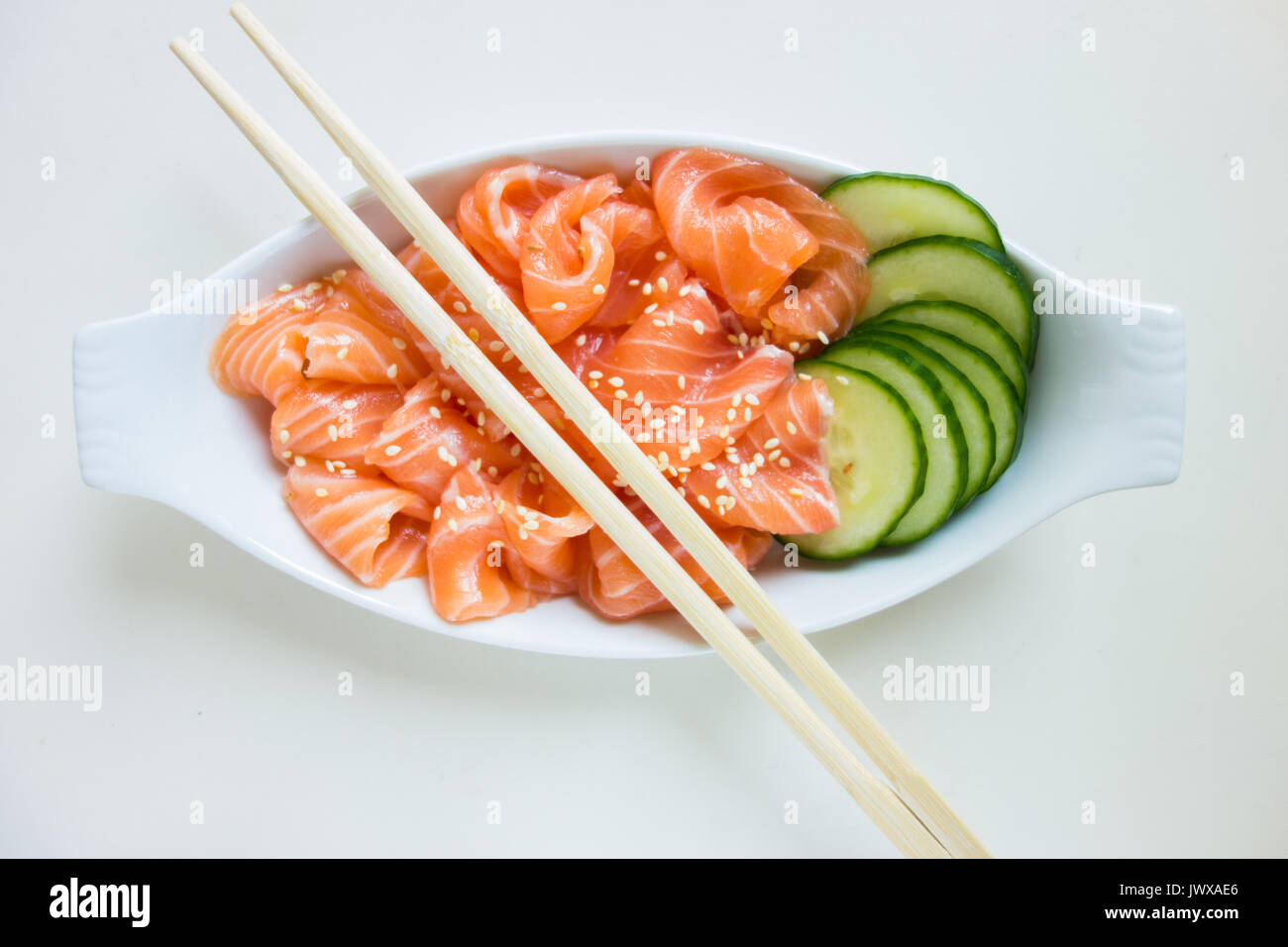 Dünne Scheiben roher Lachs Sashimi und Gurke in einer weißen Schüssel auf den weißen Hintergrund, beträufelt mit Sesam. Stockfoto