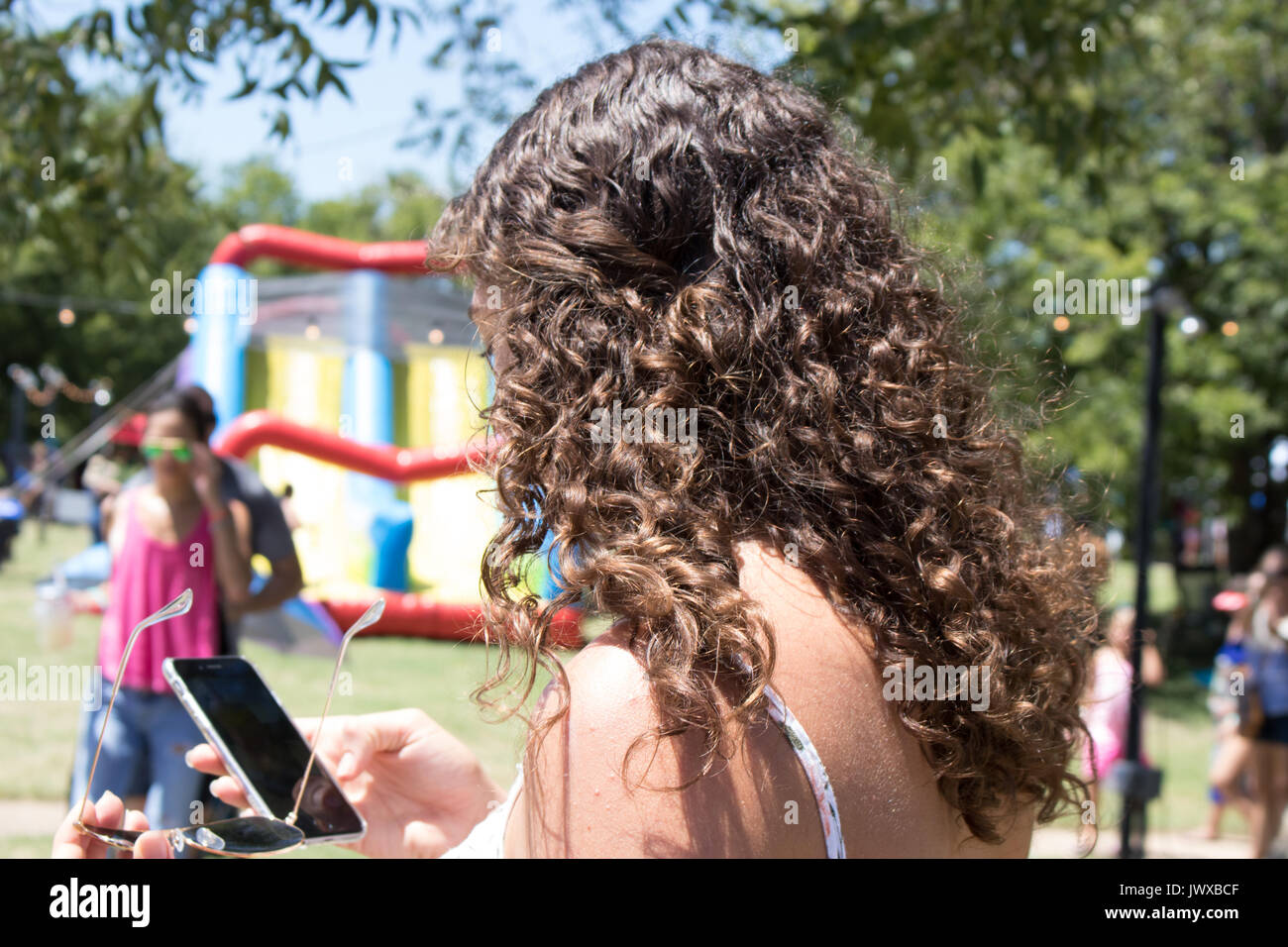 Junge weibliche Erwachsene mit braunen lockigen Haar holding Handy. Ansicht von hinter dem linken Schulter Gesicht hauptsächlich verdeckt werden. Hintergrund verschwommen Outdoor Event. Stockfoto