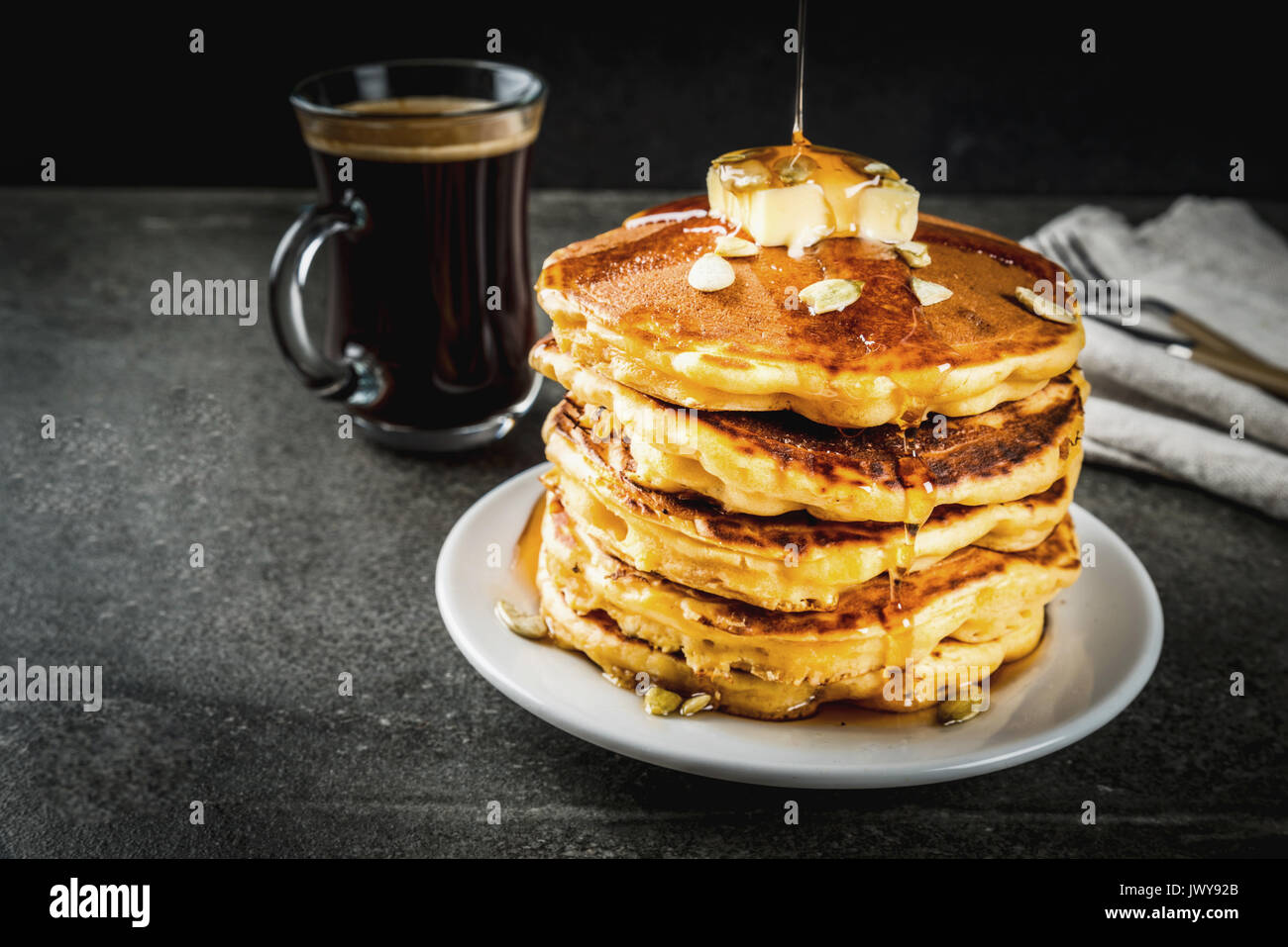 Herbstliche traditionelles Essen. Stapel von Kürbis Pfannkuchen mit Butter, Kürbiskerne und Ahornsirup. Mit einer Tasse Kaffee. Auf einem schwarzen Steintisch. Kopieren Sie sp Stockfoto